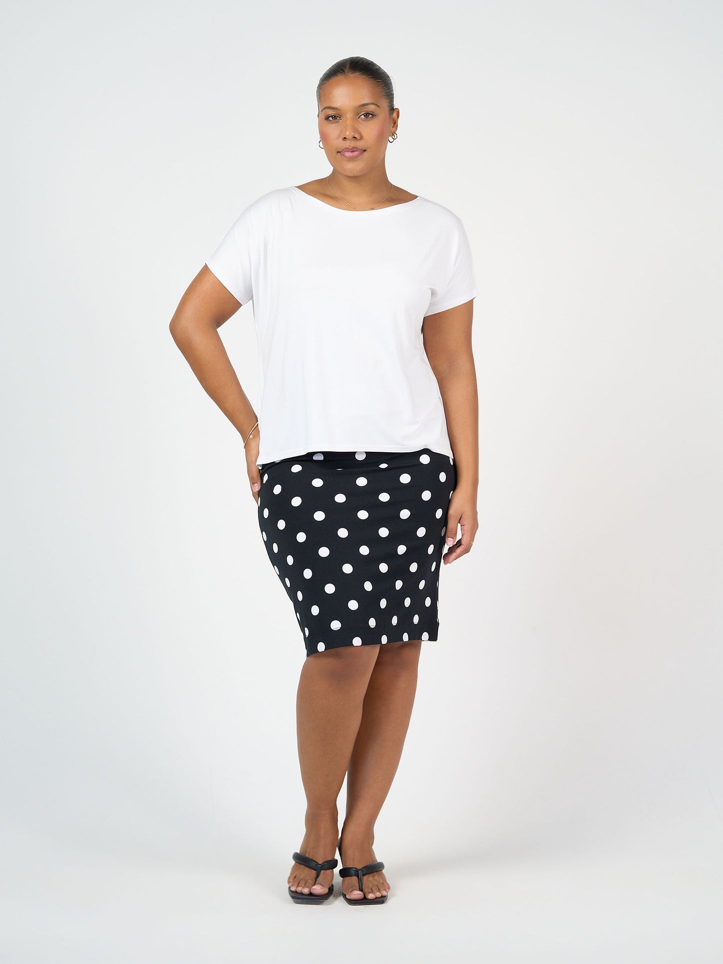 A woman poses with one hand on her hip, wearing a white short-sleeve top, black sandals, and the Midi Bamboo Whitney Tube Skirt - Polka Dot against a plain white background. She has a neutral expression.