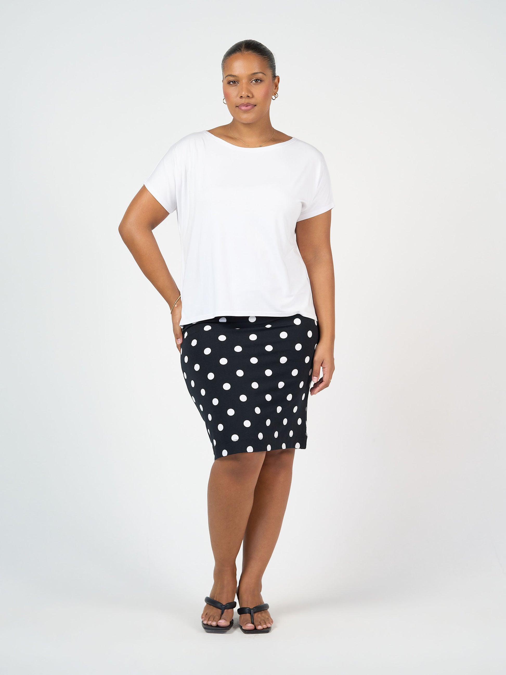 A woman poses with one hand on her hip, wearing a white short-sleeve top, black sandals, and the Midi Bamboo Whitney Tube Skirt - Polka Dot against a plain white background. She has a neutral expression.