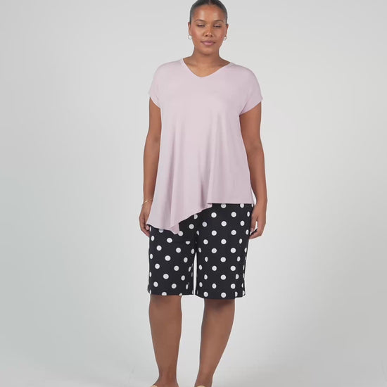 A woman stands against a plain background wearing the Holiday Asymmetrical Bamboo V-Neck T-Shirt in lilac, black knee-length shorts with white polka dots, tan slide sandals, and hoop earrings.