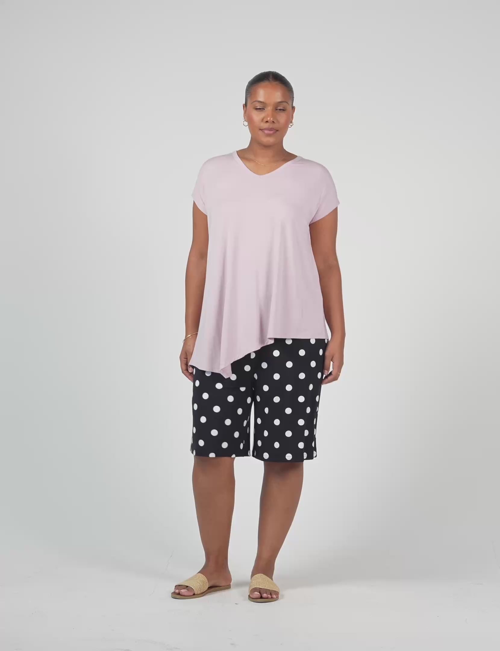 A woman stands against a plain background wearing the Holiday Asymmetrical Bamboo V-Neck T-Shirt in lilac, black knee-length shorts with white polka dots, tan slide sandals, and hoop earrings.