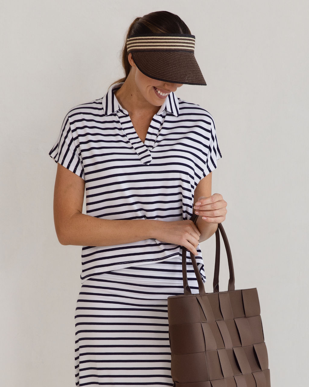A woman smiles while holding a brown woven tote bag, wearing the Rush Bamboo Short Sleeve Collared Polo Top and a brown visor with white stripes, standing against a plain light background.