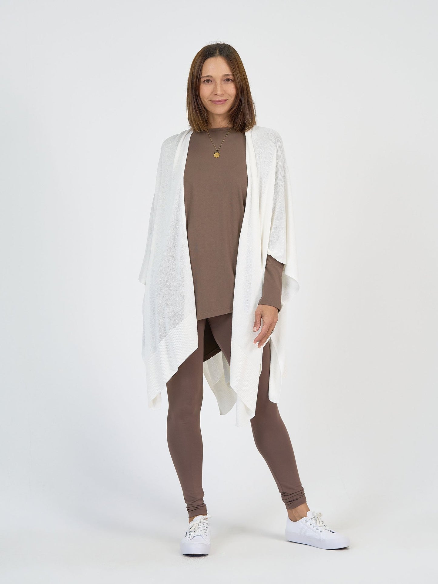 A woman faces the camera in The Cabin Set—brown long-sleeve top and leggings—with a white draped shawl and white sneakers, creating an effortlessly chic resort outfit perfect for travel, all set against a plain white background.