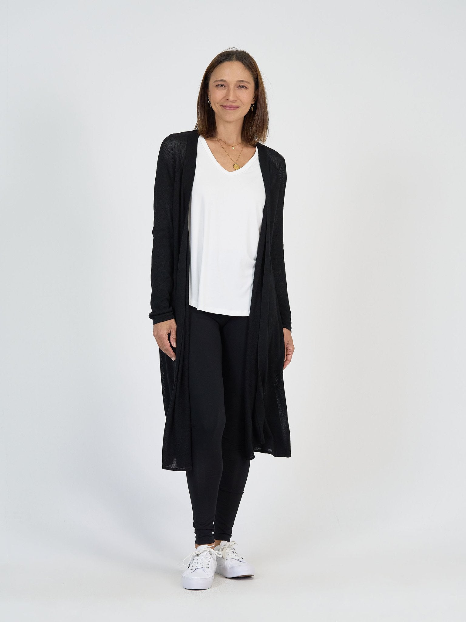 A woman with shoulder-length brown hair smiles against a white background, wearing The Alpine Set: a long black cardigan, white V-neck top, and black leggings paired with white sneakers.