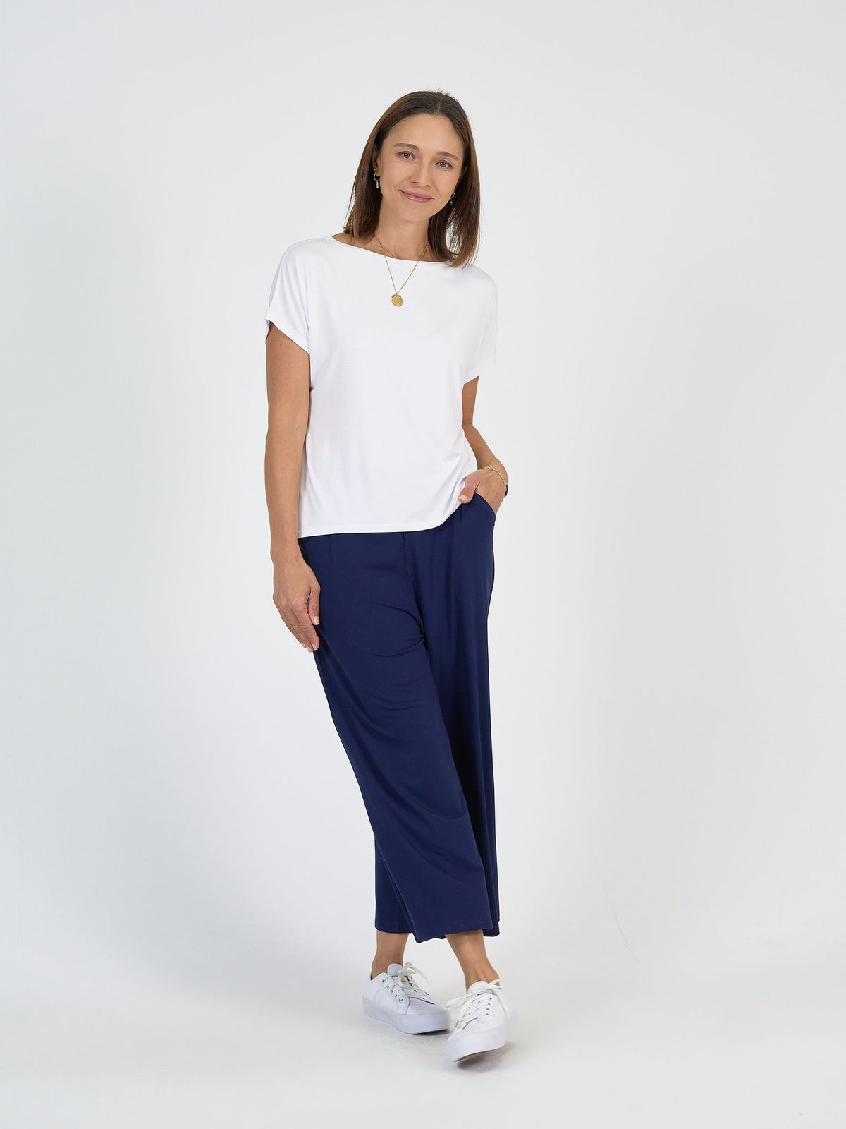 A woman with straight brown hair and a slight smile models The Sea Day Set against a white background, pairing the white short-sleeve top and loose navy blue pants with white sneakers and a gold pendant necklace. One hand rests in her pocket.