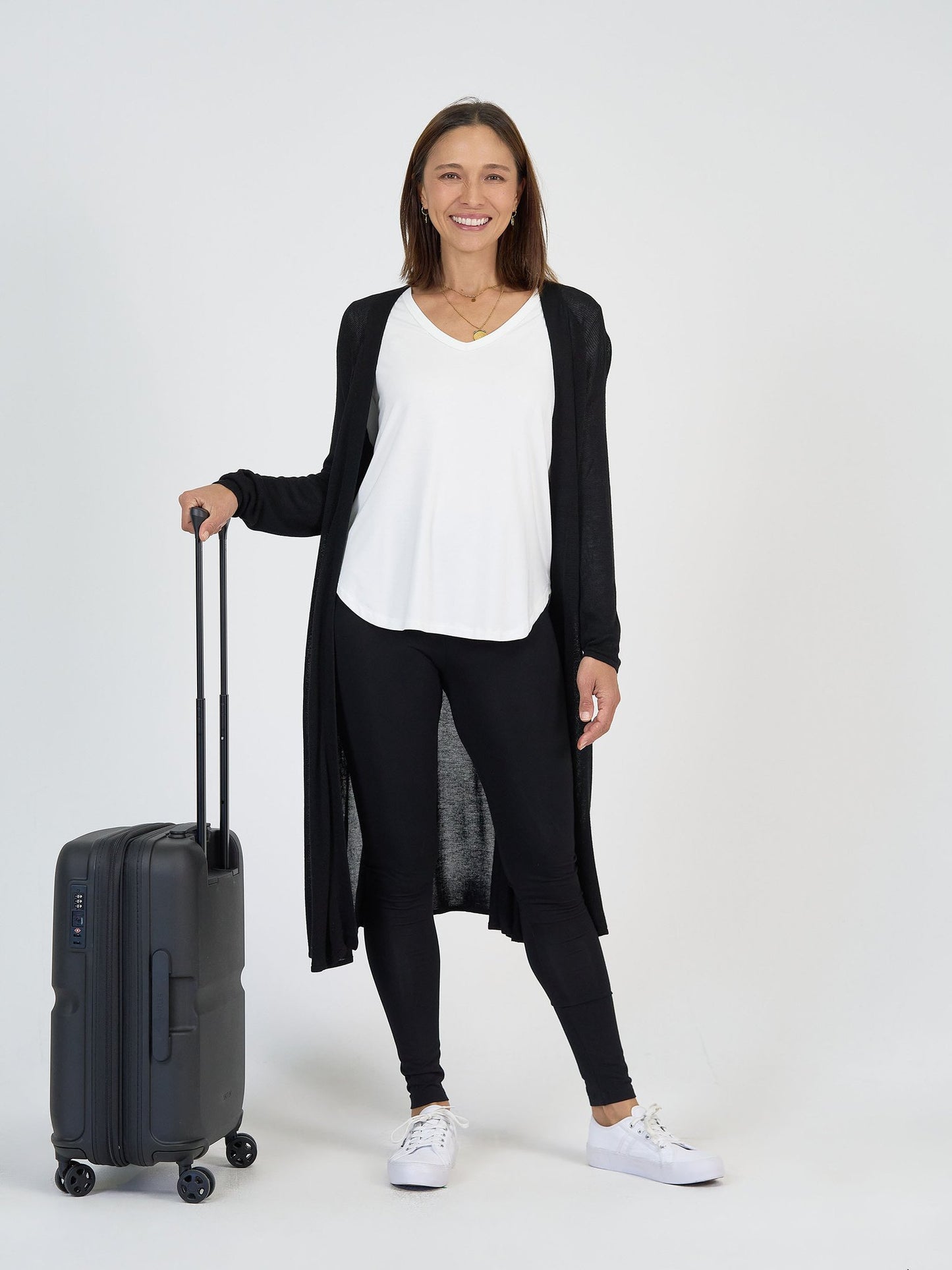 A woman stands and smiles against a white background, wearing The Alpine Set—a long black cardigan, white top, and black leggings—with white sneakers, holding a black rolling suitcase.