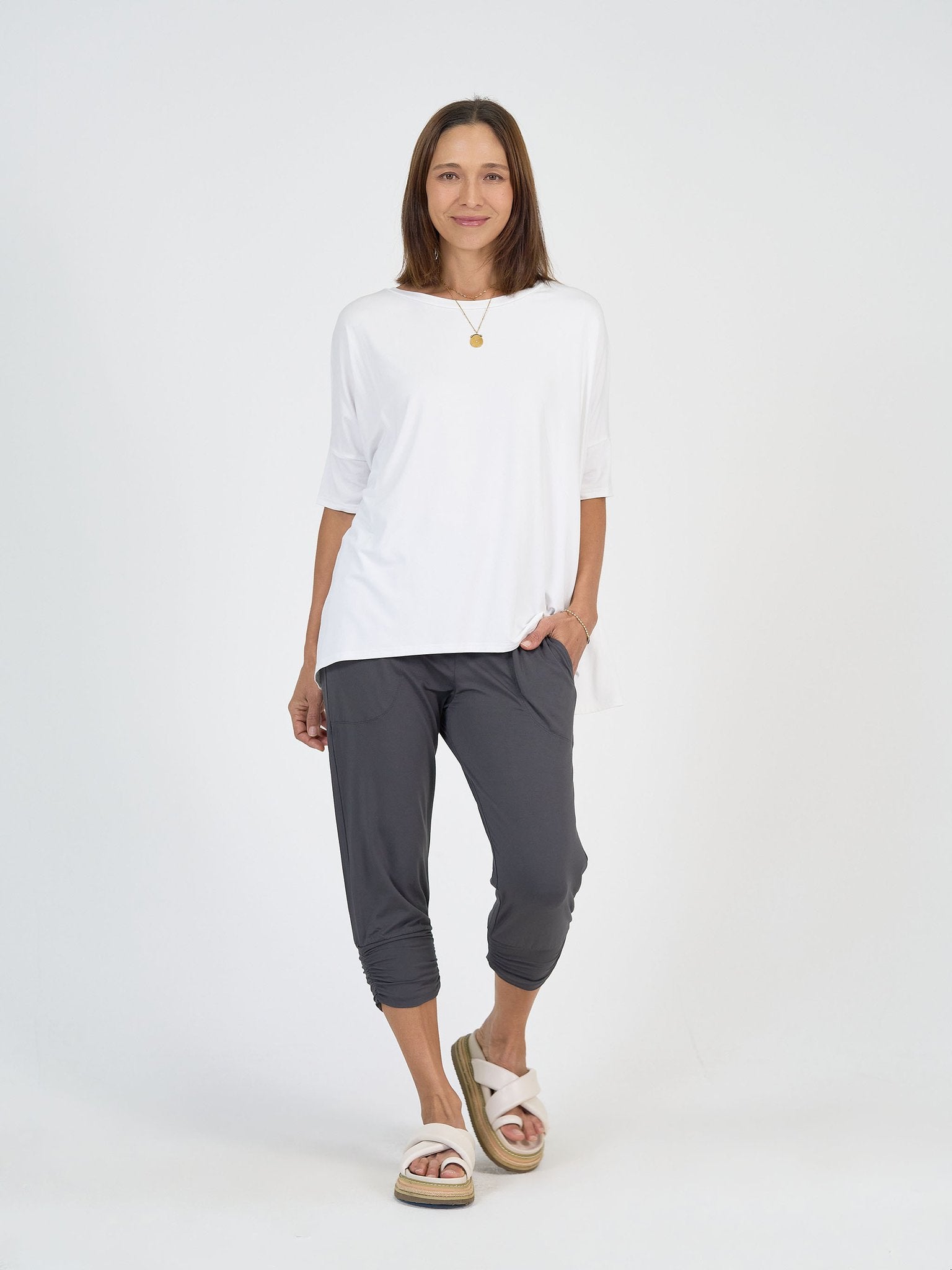 A woman stands against a white background wearing The Long Weekend Set: a loose white T-shirt and gray cropped pants, paired with white slide sandals and a gold pendant necklace, her left hand in her pocket and expression relaxed.