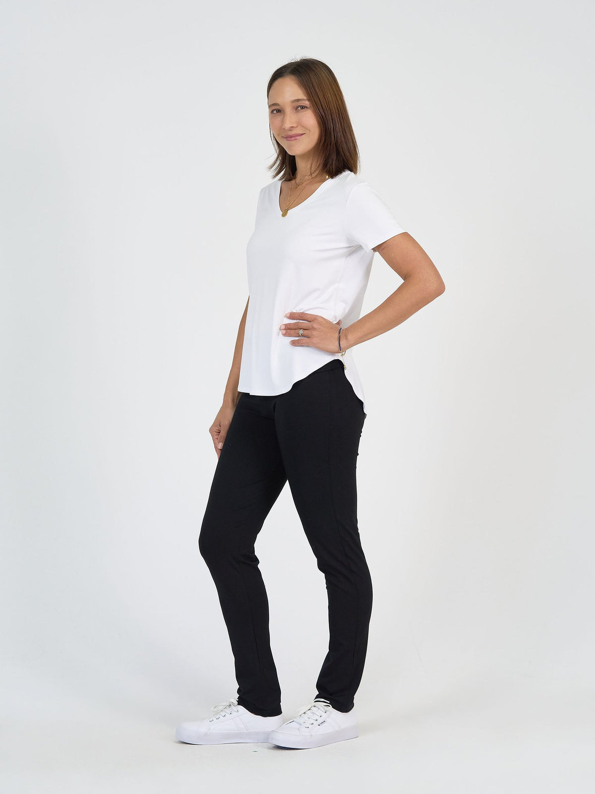 A woman stands against a plain white background, smiling, wearing The Long Haul Set—which includes a white short-sleeve V-neck top and black pants—with white sneakers. One hand is on her hip and the other rests by her side.