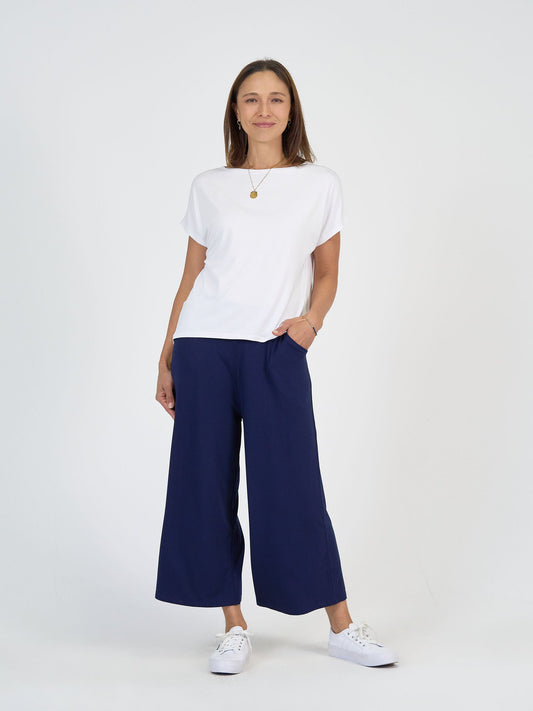 A woman with straight brown hair and a gold necklace stands against a white background, smiling softly with one hand in her pocket, wearing The Sea Day Set—featuring a white t-shirt, navy wide-leg pants, and white sneakers.