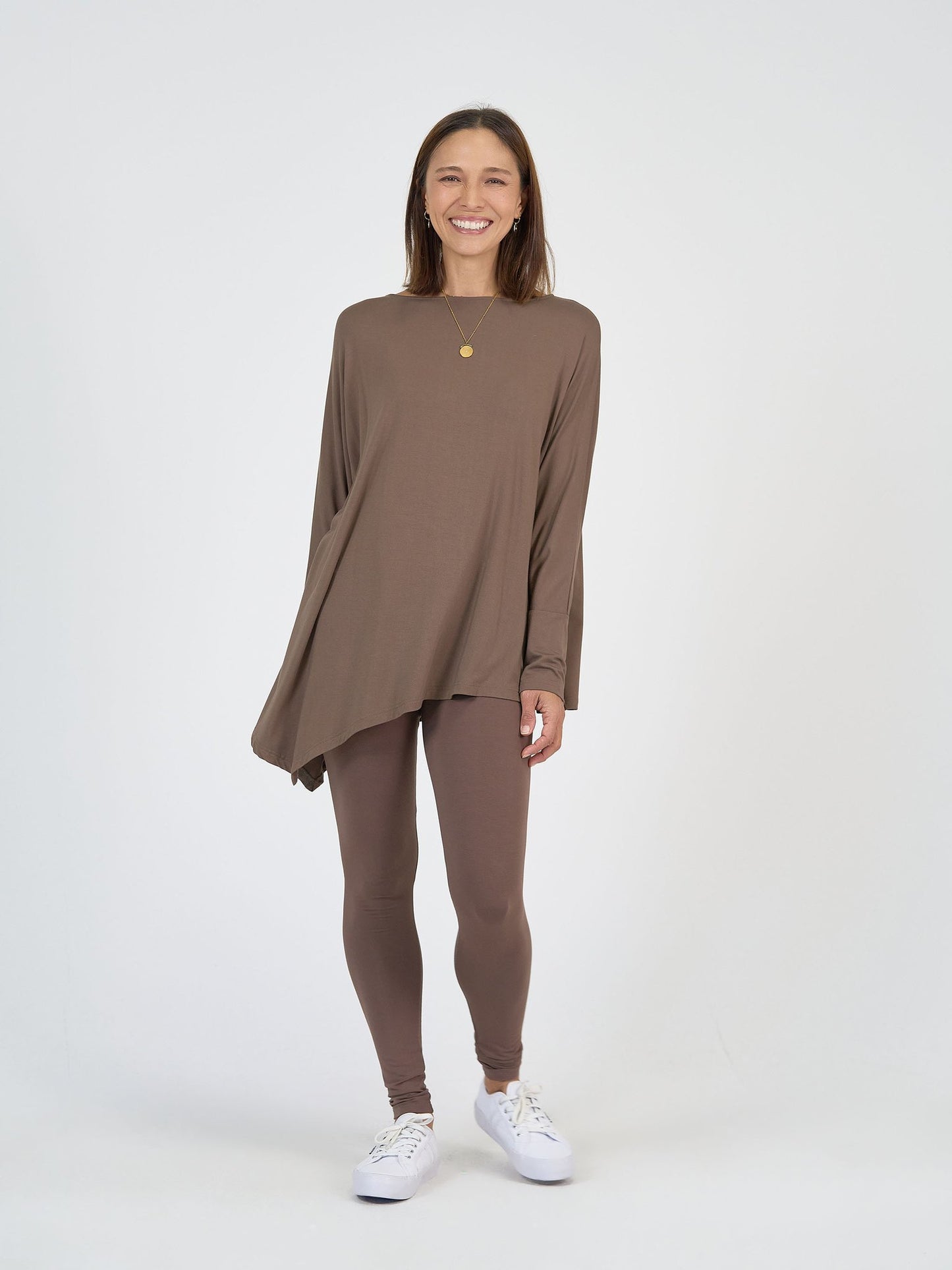 A smiling woman stands against a plain white background, wearing The Cabin Set—a loose, asymmetrical brown top with matching leggings—paired with white sneakers and a gold pendant necklace.