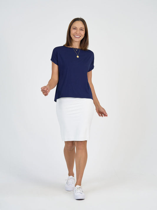 A woman smiles while walking against a plain white background, wearing The Tropics Set: a navy blue short-sleeve top, white knee-length skirt, paired with white sneakers and a gold necklace.