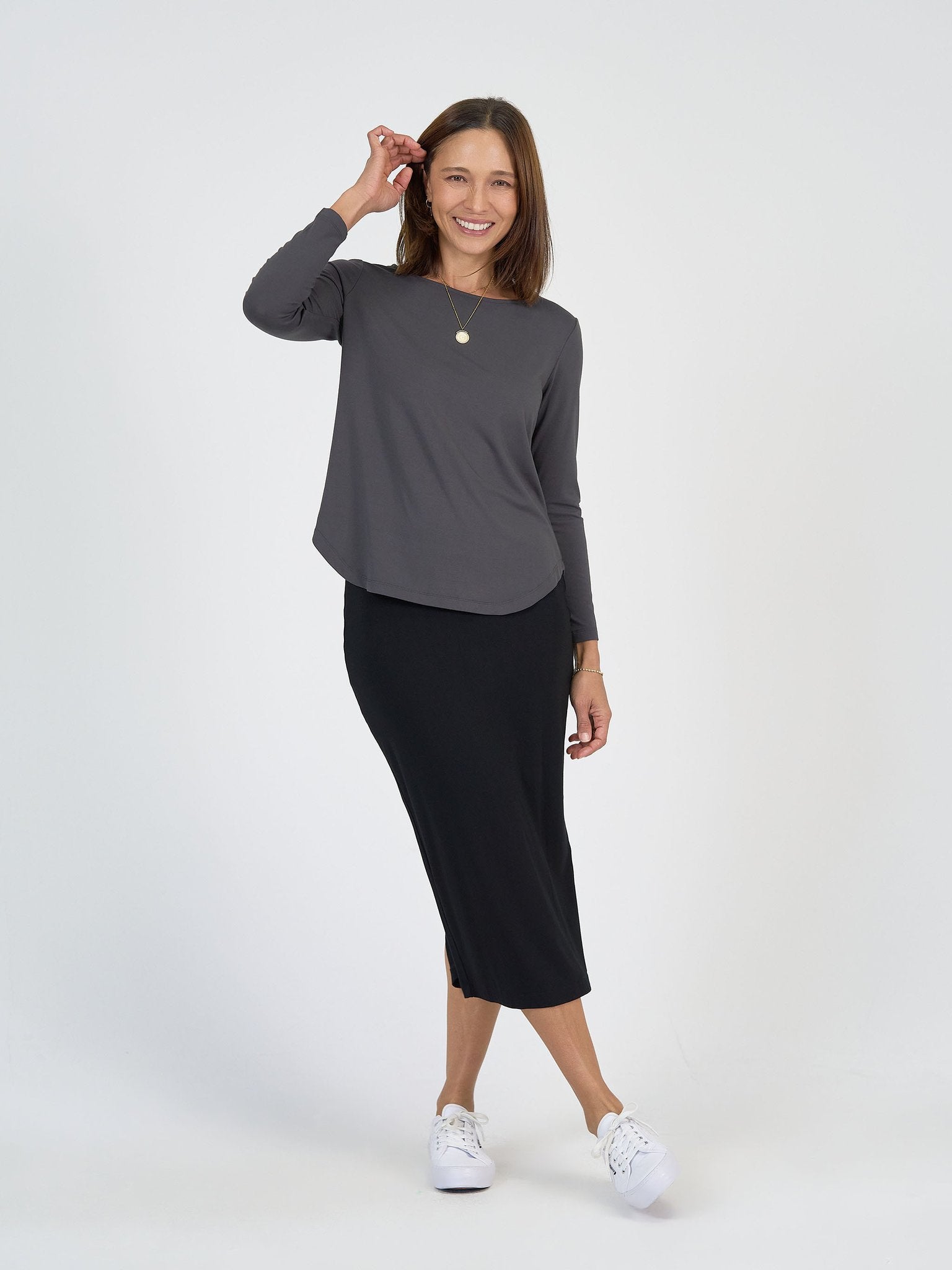 A woman smiles against a white background, wearing The Winter Sun Set—featuring a long-sleeve gray top and black midi skirt—with white sneakers, a simple necklace, and one hand near her head.