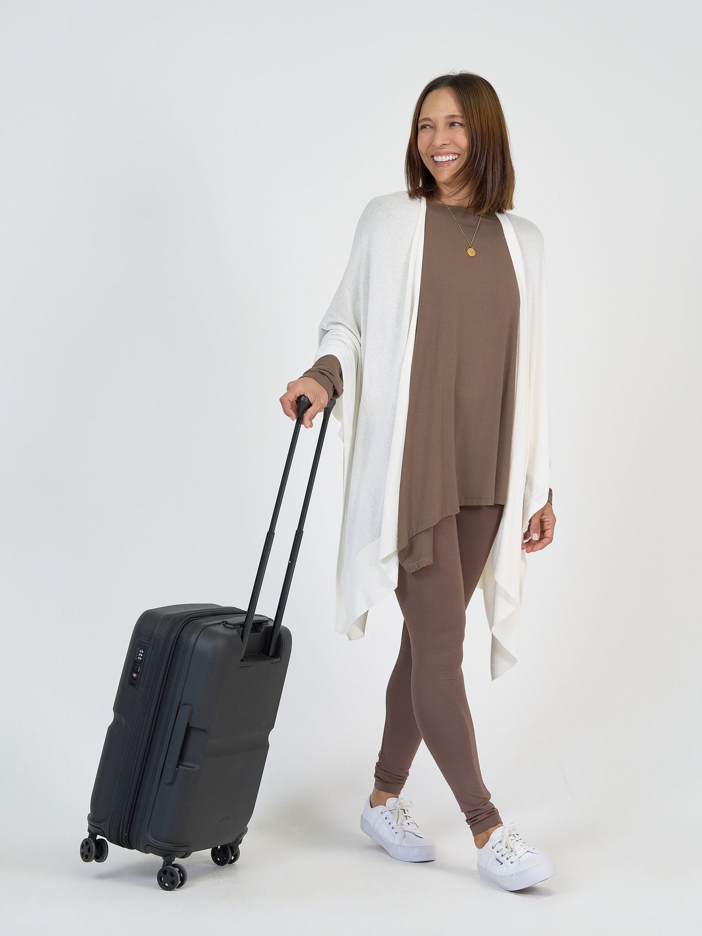 A woman smiles while pulling The Cabin Set, a black rolling suitcase. She wears a brown matching top and leggings, a white open cardigan, and white sneakers—ideal for effortless, packable resort outfits.