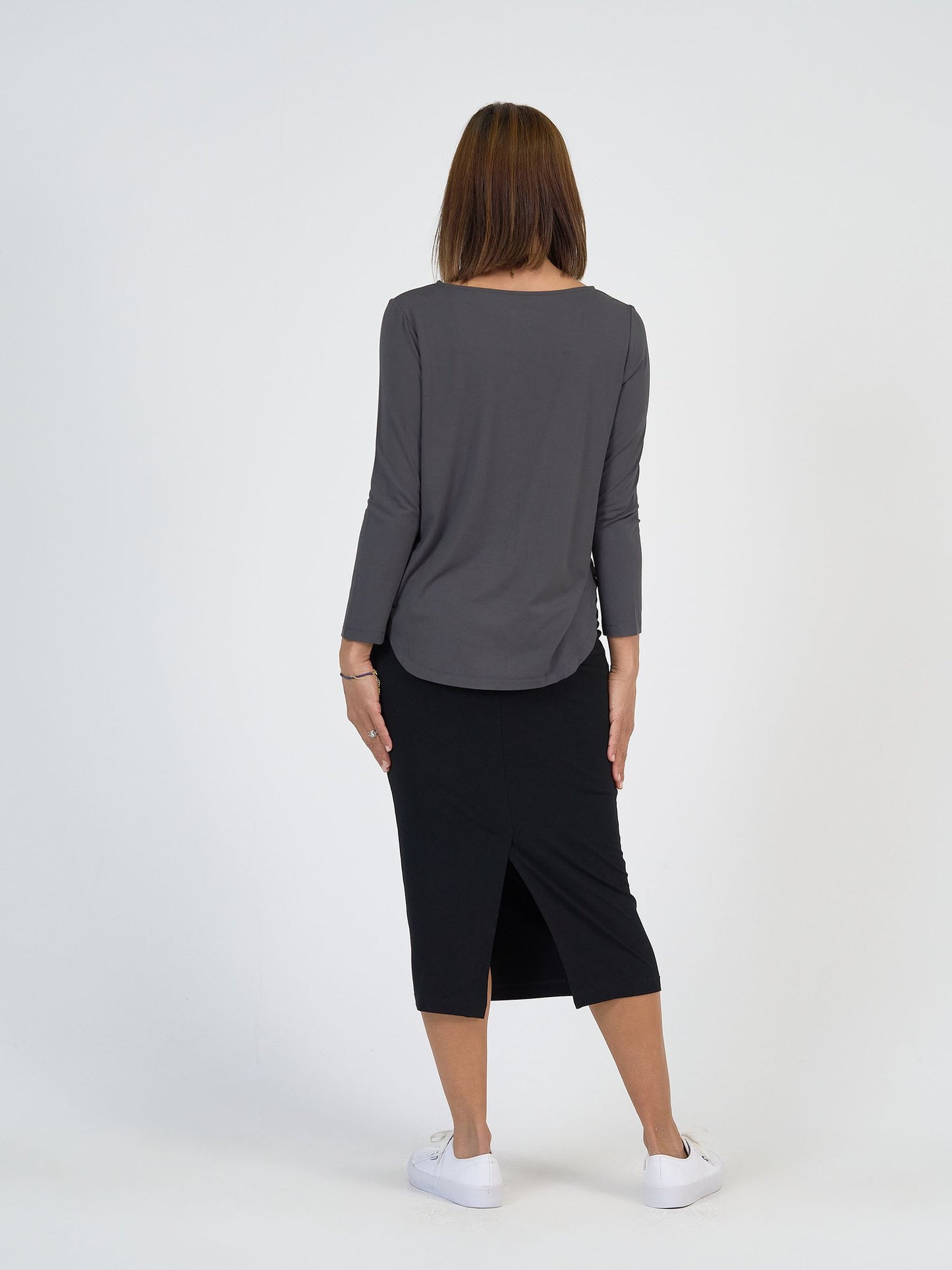 A person with straight, shoulder-length brown hair stands facing away, modeling The Winter Sun Set: a long-sleeved gray top and black knee-length skirt with back slit, paired with white sneakers against a plain white background.