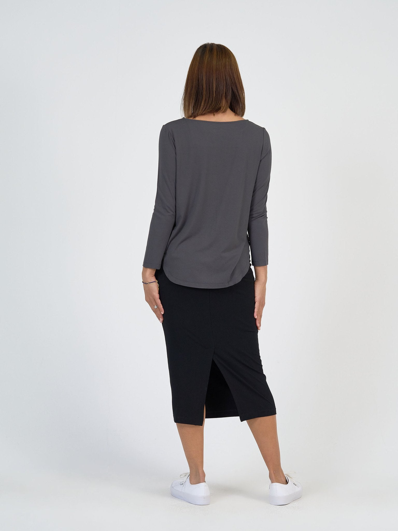 A person with straight, shoulder-length brown hair stands facing away, modeling The Winter Sun Set: a long-sleeved gray top and black knee-length skirt with back slit, paired with white sneakers against a plain white background.
