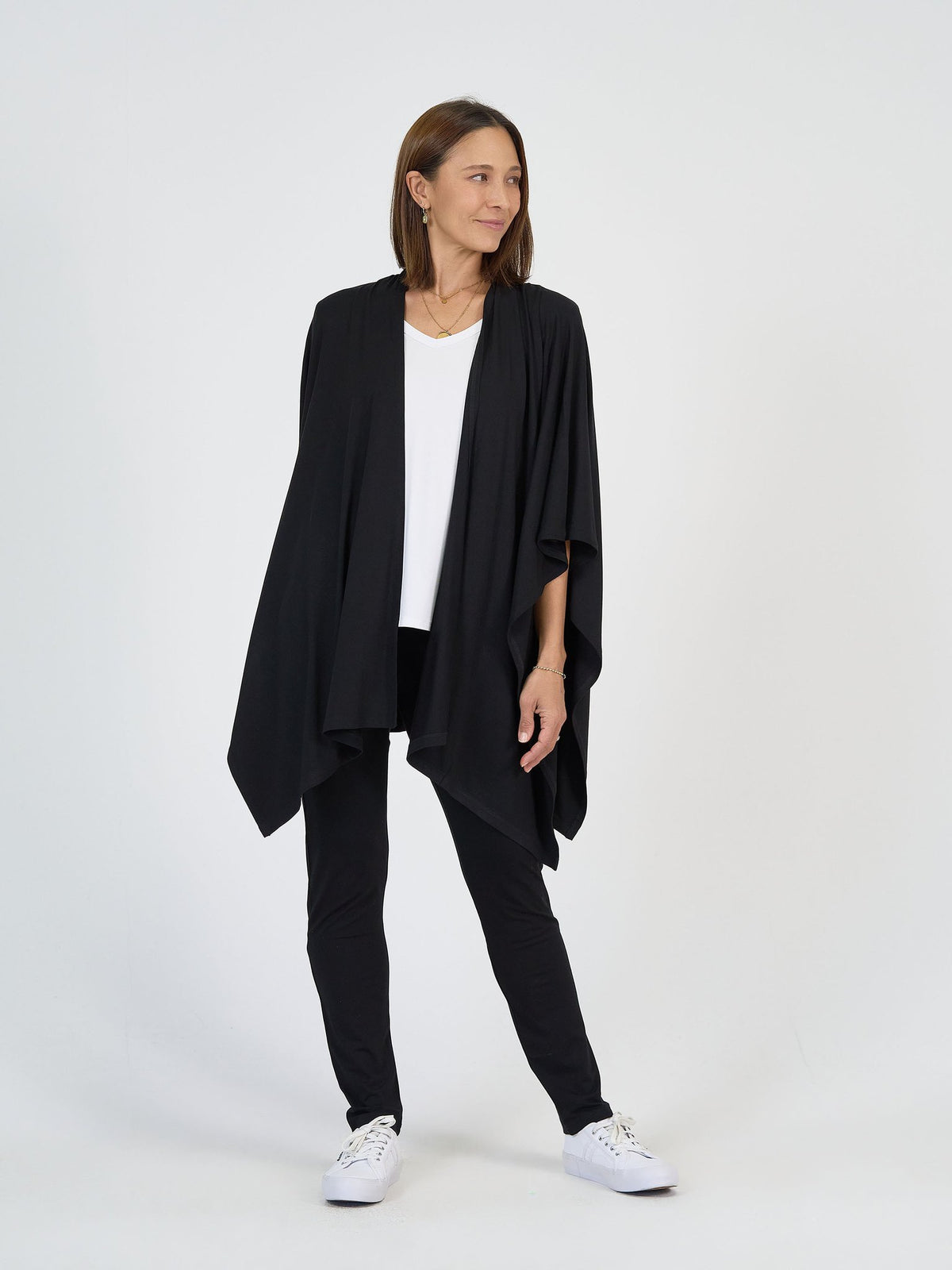 A woman wears The Long Haul Set—black draped cardigan, white top, black pants—paired with white sneakers against a plain white background. She looks to her left and smiles slightly.