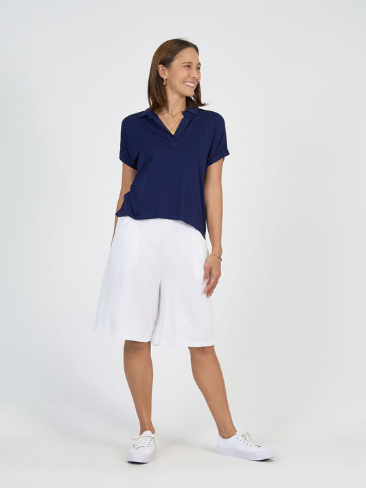 A woman poses with a smile against a white background, wearing The Mediterranean Set—a navy short-sleeve polo shirt and white knee-length shorts—with white sneakers. She has short brown hair and keeps one hand in her pocket.