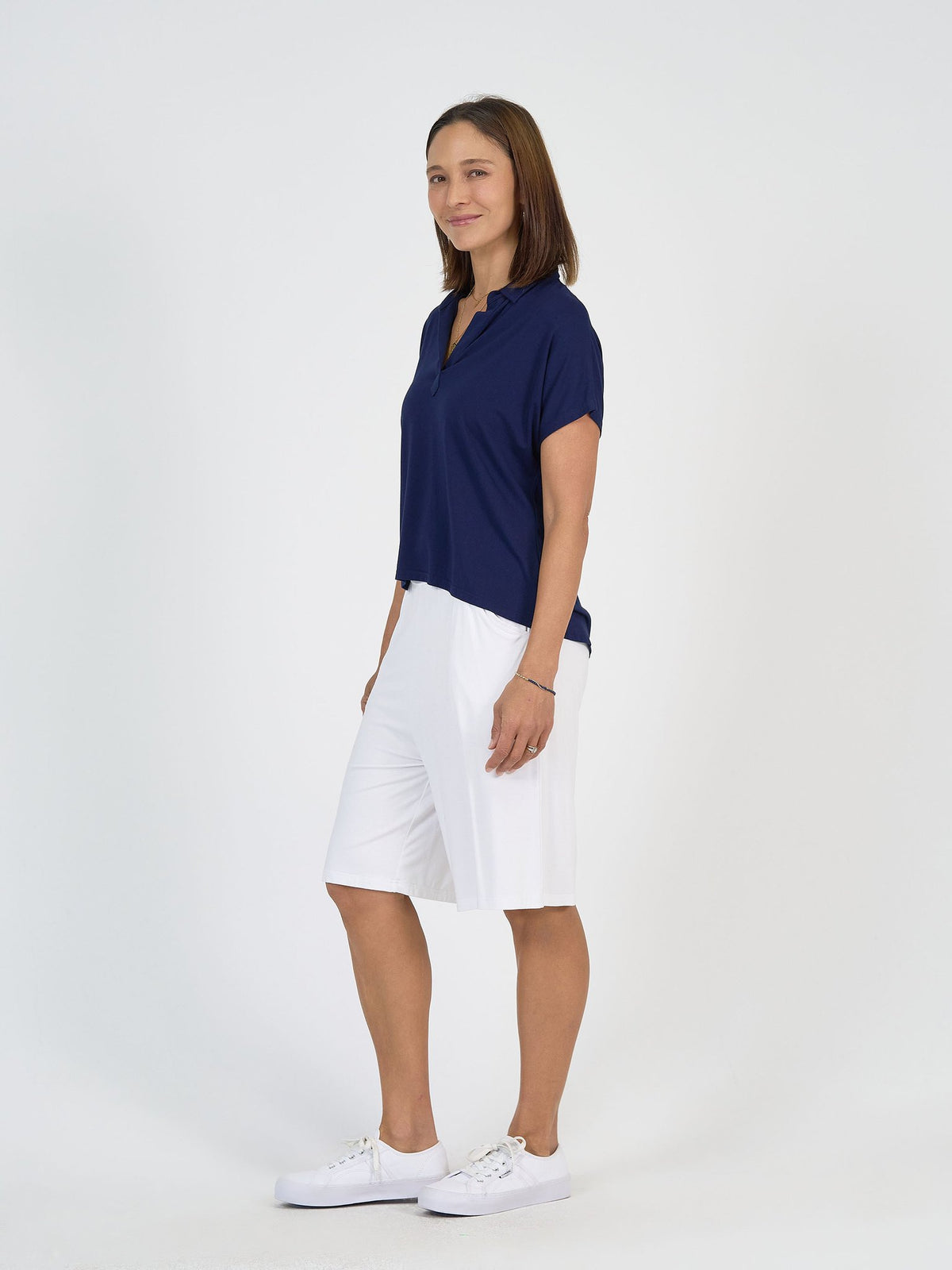 A woman stands sideways, smiling, wearing The Mediterranean Set—a navy short-sleeve polo shirt and white knee-length shorts—with white sneakers against a plain white background.