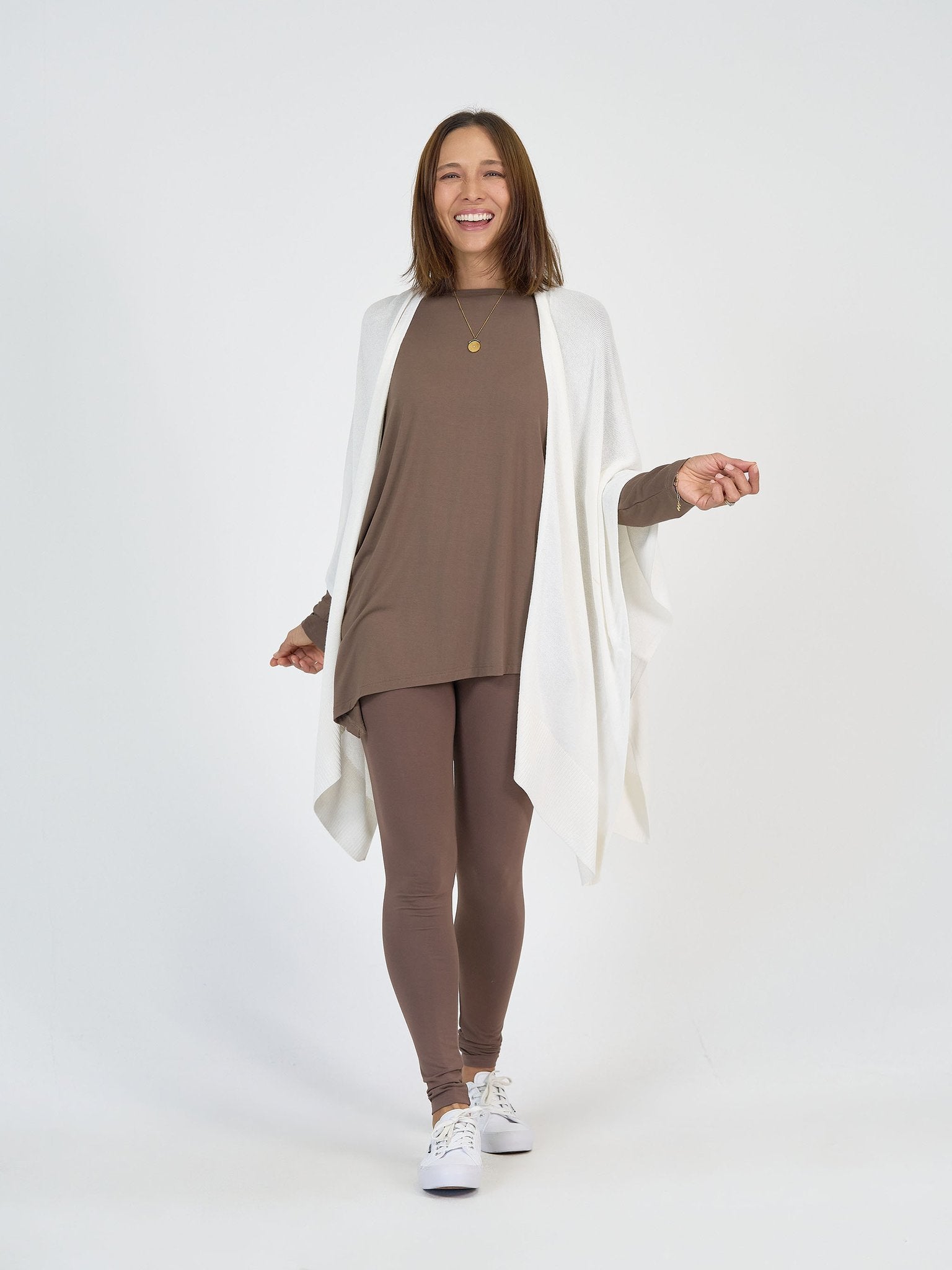 A woman smiles against a white background, wearing The Cabin Set—a loose brown tunic with matching leggings—perfect as resort wear and packable travel clothing, styled with a white shawl, sneakers, and a gold necklace.