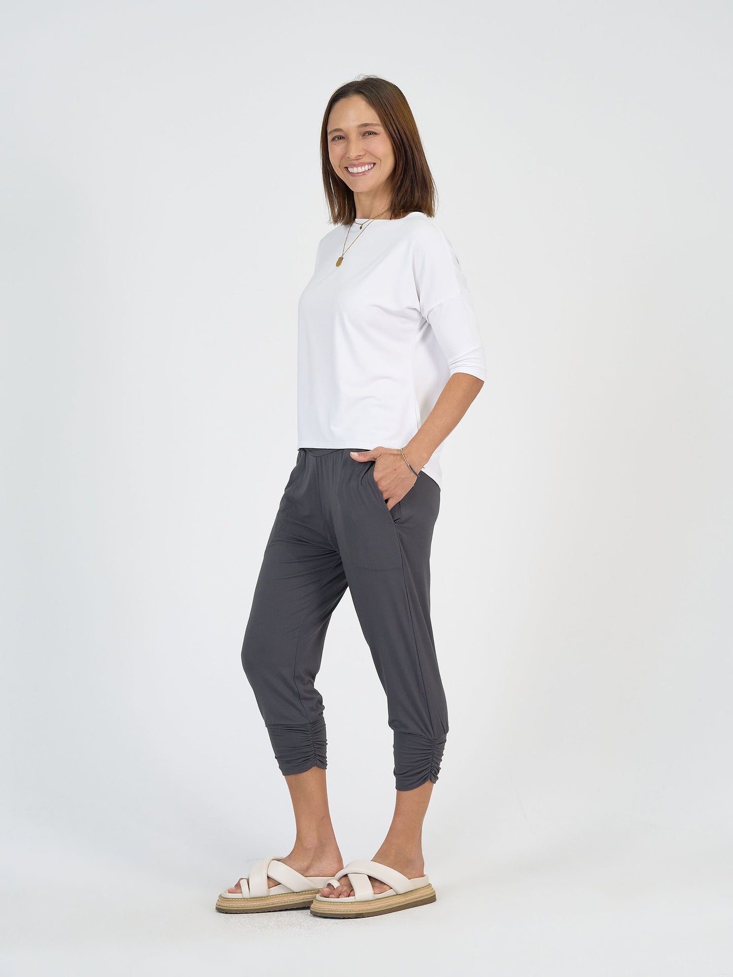 A woman with shoulder-length brown hair smiles against a white background, wearing The Long Weekend Set—featuring a white top and gray cropped pants with pockets—paired with white platform sandals. Her hands rest in her pockets.