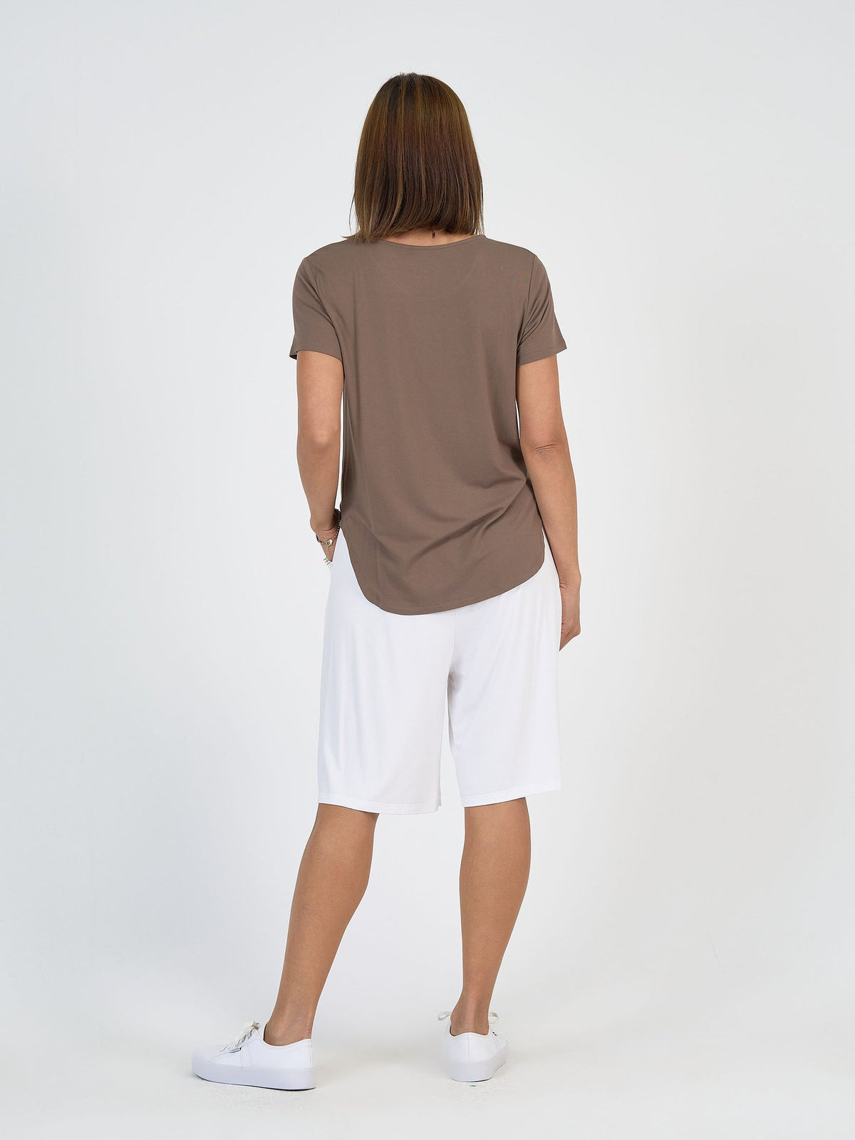 A person stands with their back to the camera, wearing The Mediterranean Set: a brown short-sleeve top and white knee-length shorts, paired with white sneakers against a plain white background.