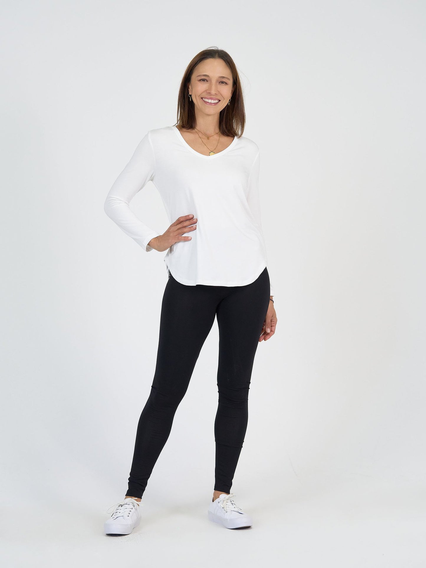 A woman stands smiling against a plain white background, wearing The Alpine Set with a long-sleeved white shirt, black leggings, and white sneakers. She has one hand on her hip and the other relaxed by her side.
