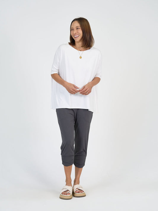 A woman smiles against a plain white background, wearing The Long Weekend Set: a loose white top and dark gray cropped pants, paired with white slide sandals and a gold necklace.