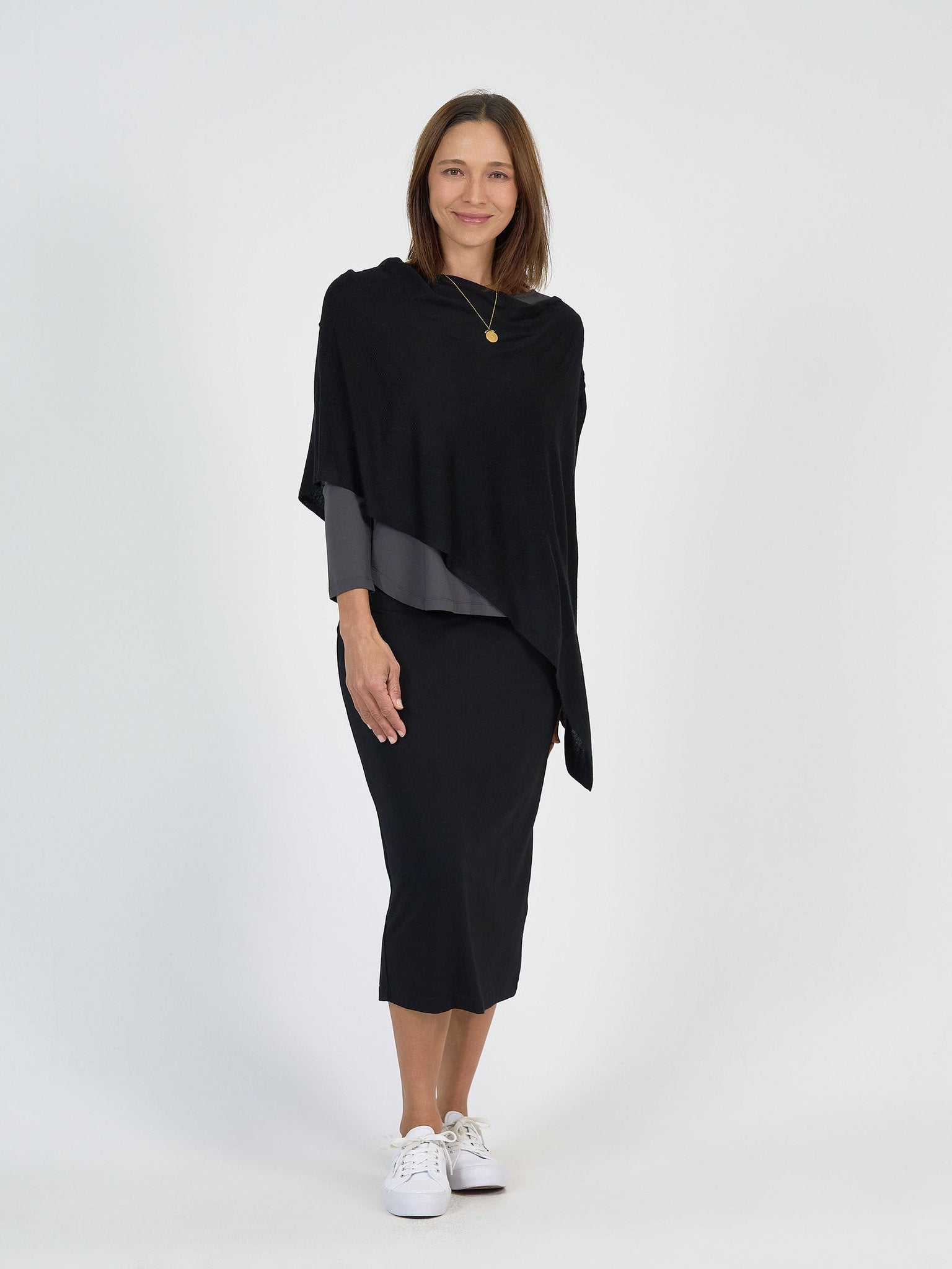 A woman with shoulder-length brown hair smiles against a white background, wearing The Winter Sun Set—a black asymmetrical poncho over a gray top and black midi skirt—paired with white sneakers and a gold pendant necklace.