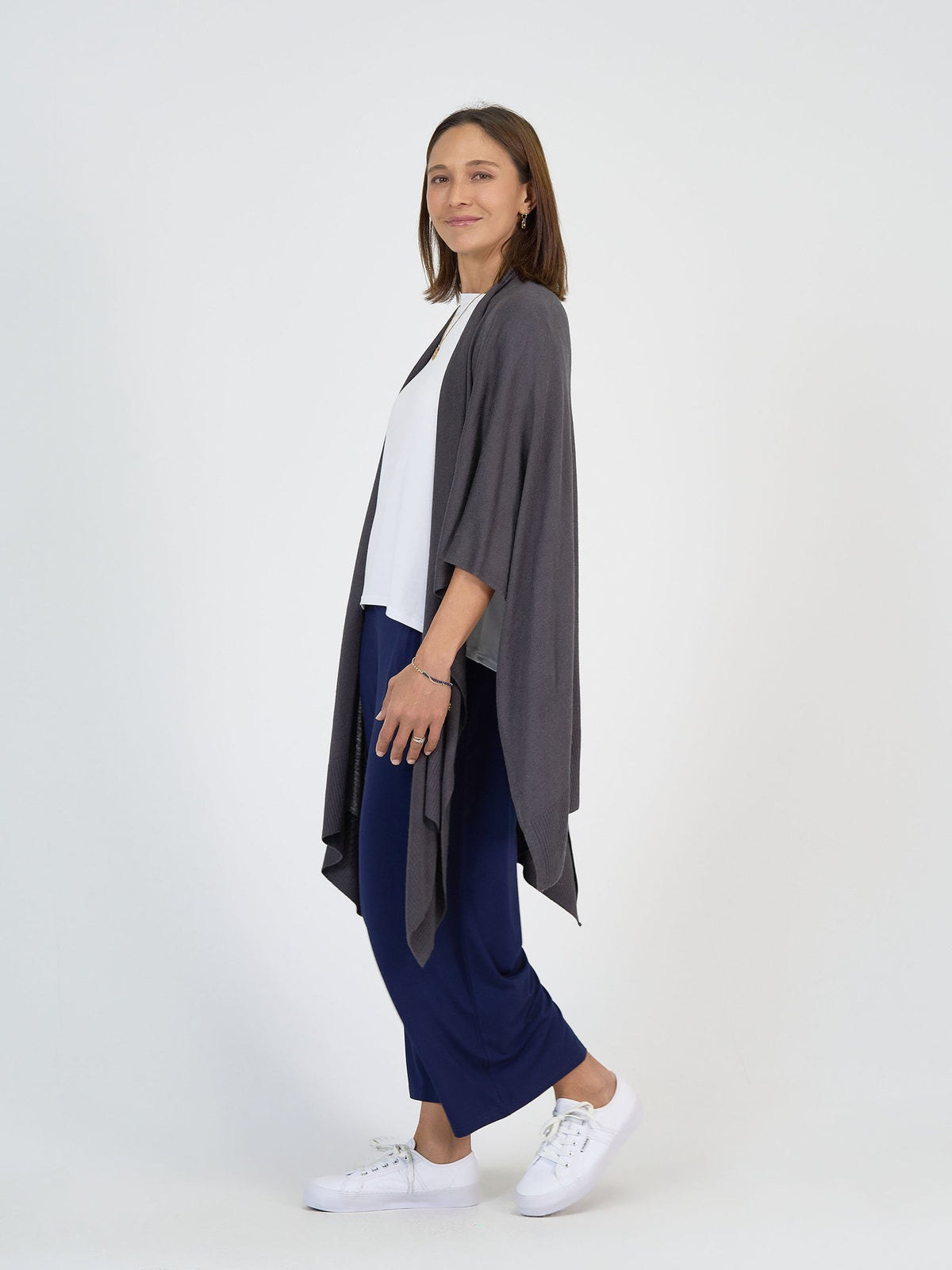 A woman smiles, standing sideways in The Sea Day Set: a loose gray cardigan over a white top and blue wide-leg pants with white sneakers, against a plain white background.