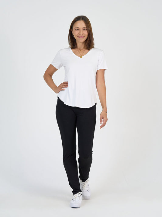 A woman smiles against a white background wearing The Long Haul Set: a white V-neck T-shirt, black pants, white sneakers, and a gold necklace. One hand is on her hip, the other rests by her side.